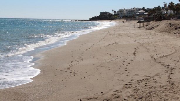 Une plage de sable avec des empreintes menant à l’eau, un ciel dégagé et des bâtiments en arrière-plan.