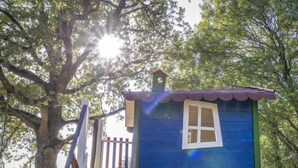 Una casita de juegos azul con una ventana blanca, techo rojo y puerta verde, rodeada de árboles y una valla de madera.
