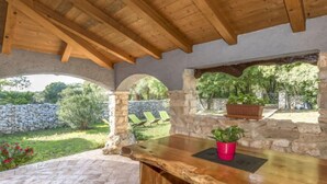 Un espacio de descanso rústico al aire libre con mesa y sillas de madera, paredes de piedra y vistas a un jardín y árboles.