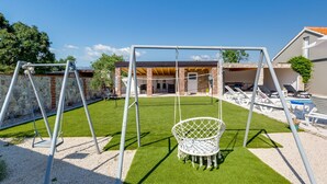 Un patio trasero con un parque infantil, césped artificial, un muro de piedra y una zona de piscina.
