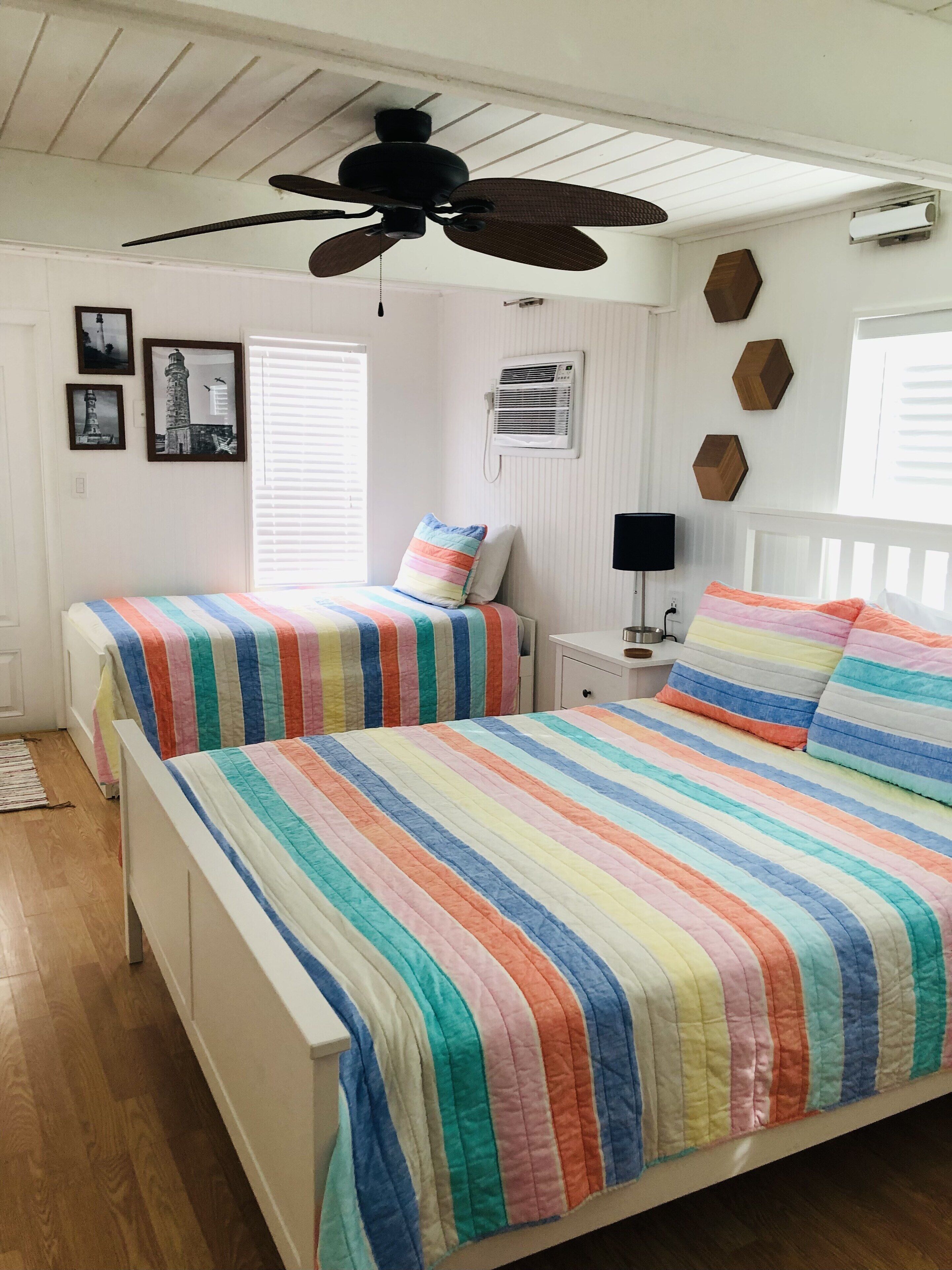 A bedroom with two beds, a ceiling fan, a window with blinds, and a wall-mounted air conditioner.