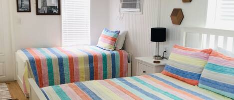 A bedroom with two beds, a ceiling fan, a window with blinds, and a wall-mounted air conditioner.