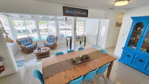 A spacious living area with a wooden table, blue chairs, wicker armchairs, and a foosball table. A sign reads "YELLOWTAIL GETAWAY BEACH HOUSE" and a blue cabinet is visible. - Yellowtail Getaway 3 bedrooms,4 beds,3 bath,boat dock,ice maker and heated pool (Marathon)