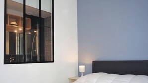 A bedroom with a bed, a nightstand, a lamp, and a large mirror reflecting a bathroom with a shower. - Ker Louis by Interhome (Dinard)