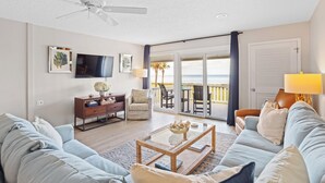 A living room with a sofa, coffee table, and TV. There is a balcony with a view of the ocean. - 621 Surf Villas, Oceanfront Condo, BAM Vacation Rentals (Ponte Vedra Beach)