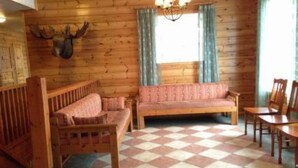 Een knus interieur van een blokhut met een geruit vloerkleed, houten wanden, een opgehangen hertenhoofd, een bank en een eettafel.