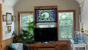 Ein gemütliches Zimmer mit Holzschreibtisch, Fernseher, Stuhl und einem großen Fenster mit Blick auf Bäume.