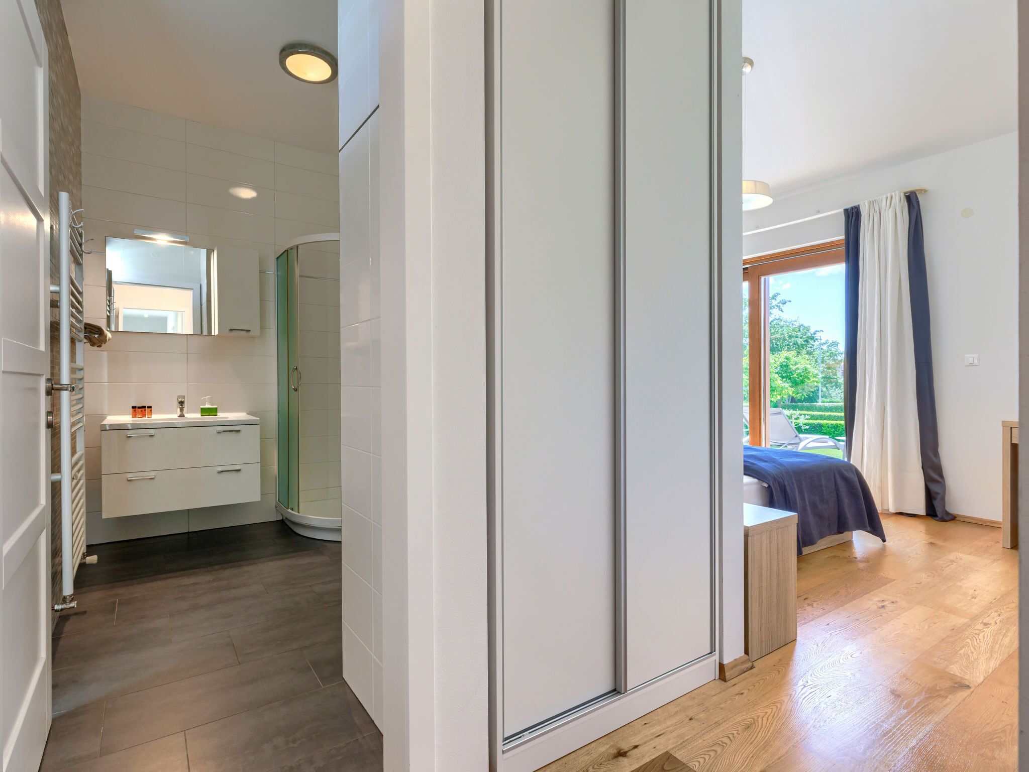 A modern bathroom with a shower, toilet, and sink. There is a large mirror above the sink and a window with curtains.
