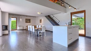 A spacious kitchen with a central island, a dining area with chairs, a staircase, and a large window with a view of greenery.