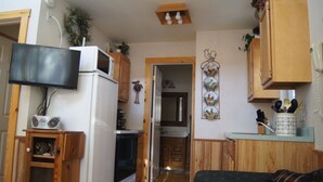Cocina compacta con gabinetes de madera, horno integrado, microondas y televisor sobre un mueble.