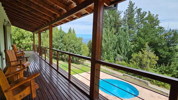 A wooden deck with chairs overlooking a swimming pool and a lush green landscape. - Buena Vida Social Club (El Bolson)