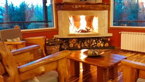 A cozy room with a stone fireplace, wooden furniture, and a view of trees through large windows. - Buena Vida Social Club (El Bolson)