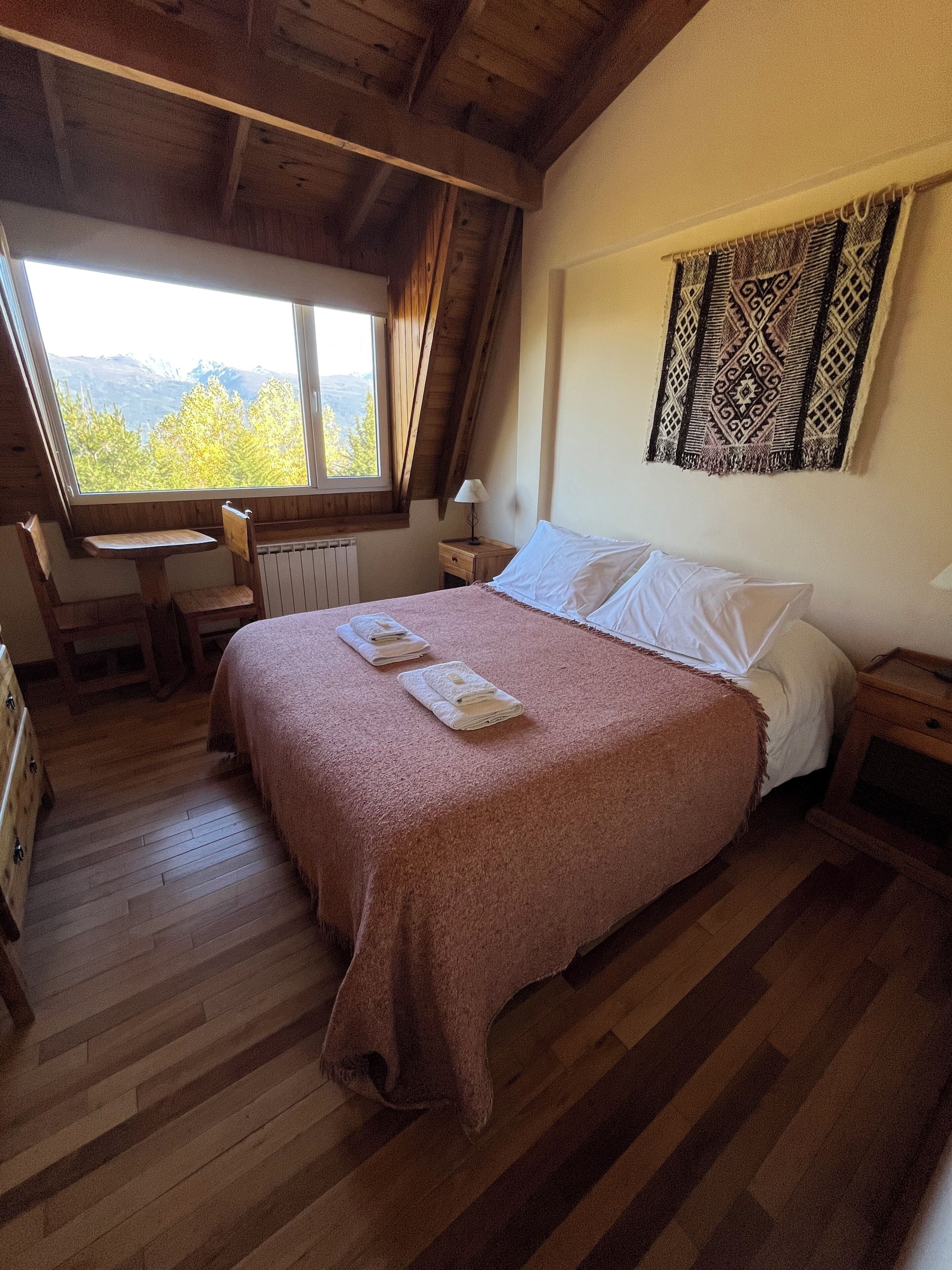 A bedroom with a wooden ceiling, a large window, a bed with white linens, a small table, and a chair.