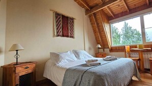 A cozy bedroom with a wooden ceiling, a large window with a view of trees, a bed with white linens, and a small desk with a chair. - Buena Vida Social Club (El Bolson)