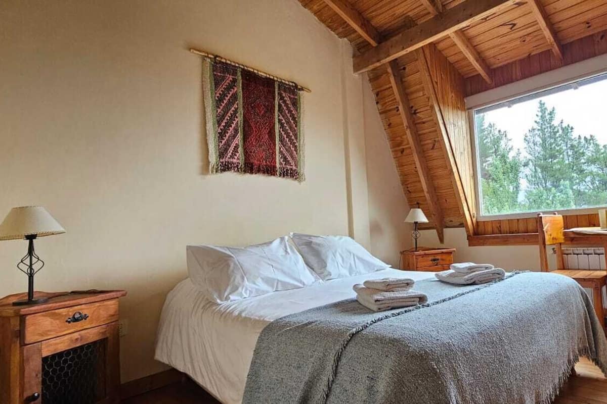 A cozy bedroom with a wooden ceiling, a large window with a view of trees, a bed with white linens, and a small desk with a chair.