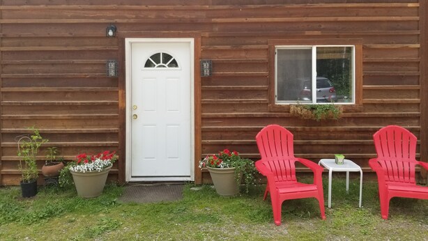 A wooden cabin with a white door, two red Adirondack chairs, a small table, and potted plants. - “Cozy Mountain Suites - #5 just 5 minutes from Glacier Park (Coram)