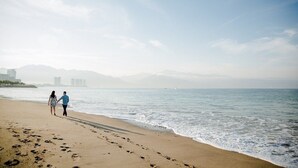 Une plage où l’on voit des empreintes de pas menant à l’eau, un couple qui se promène et des bâtiments qui se dressent au loin.