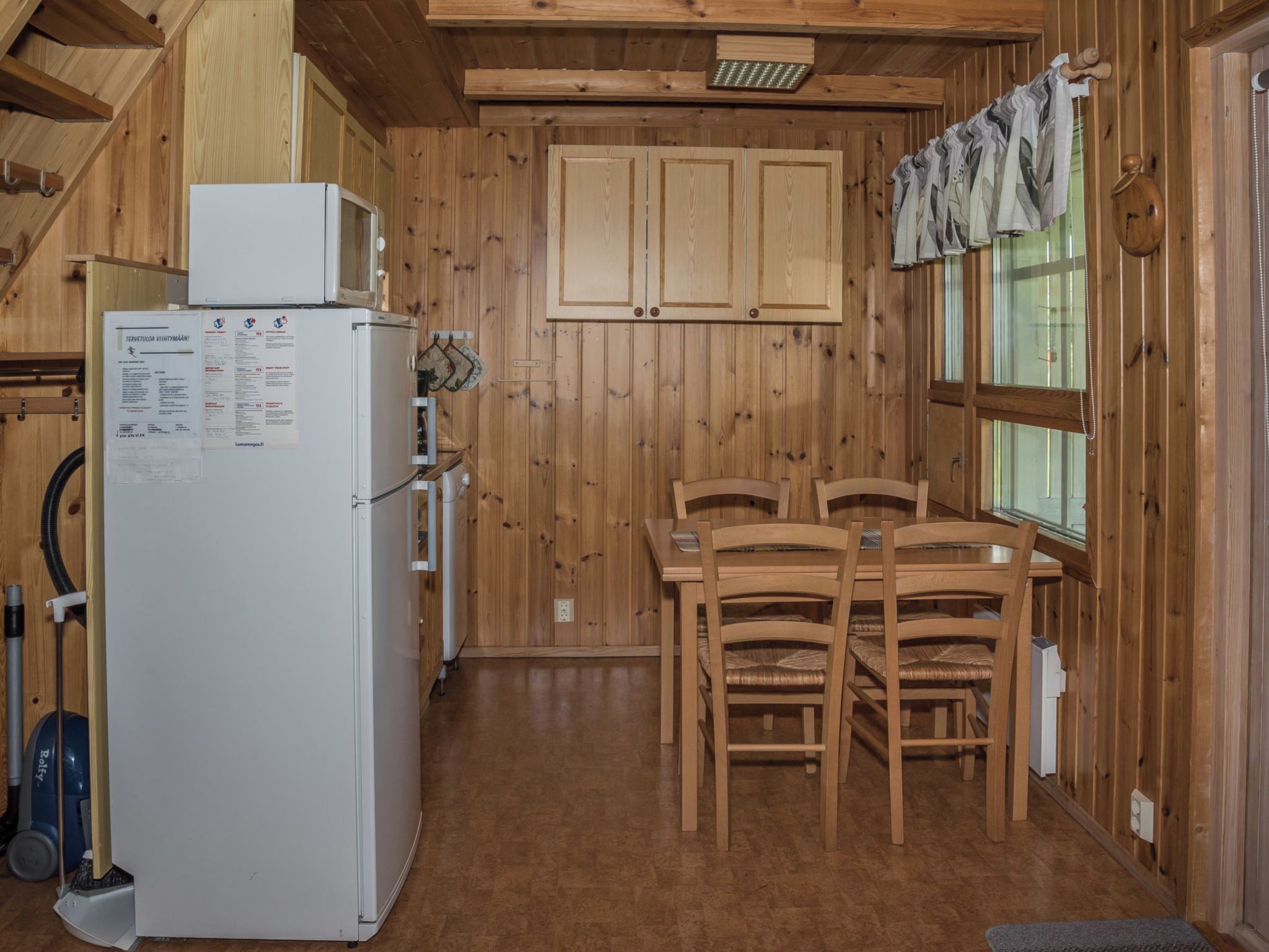 A wooden cabin interior with a refrigerator, wooden cabinets, a dining table with chairs, and a window with curtains.