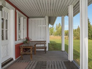 Un porche abrité avec un banc et une table en bois, une porte rouge et une vue sur un champ verdoyant.