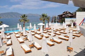 Un área de descanso en la playa con tumbonas y sombrillas, con vista a una piscina y al mar.