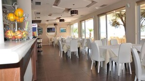A dining area with white chairs and tables, a fruit display, and a Bull sign. - Hotel Coral (Vlorë)