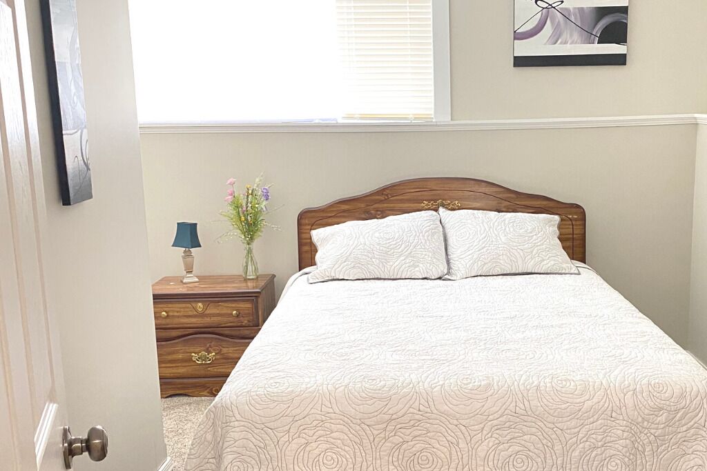 Een slaapkamer met een houten hoofdbord, een bed met een geborduurd beddegoed, een nachtkastje met een lamp en een vaas met bloemen.
