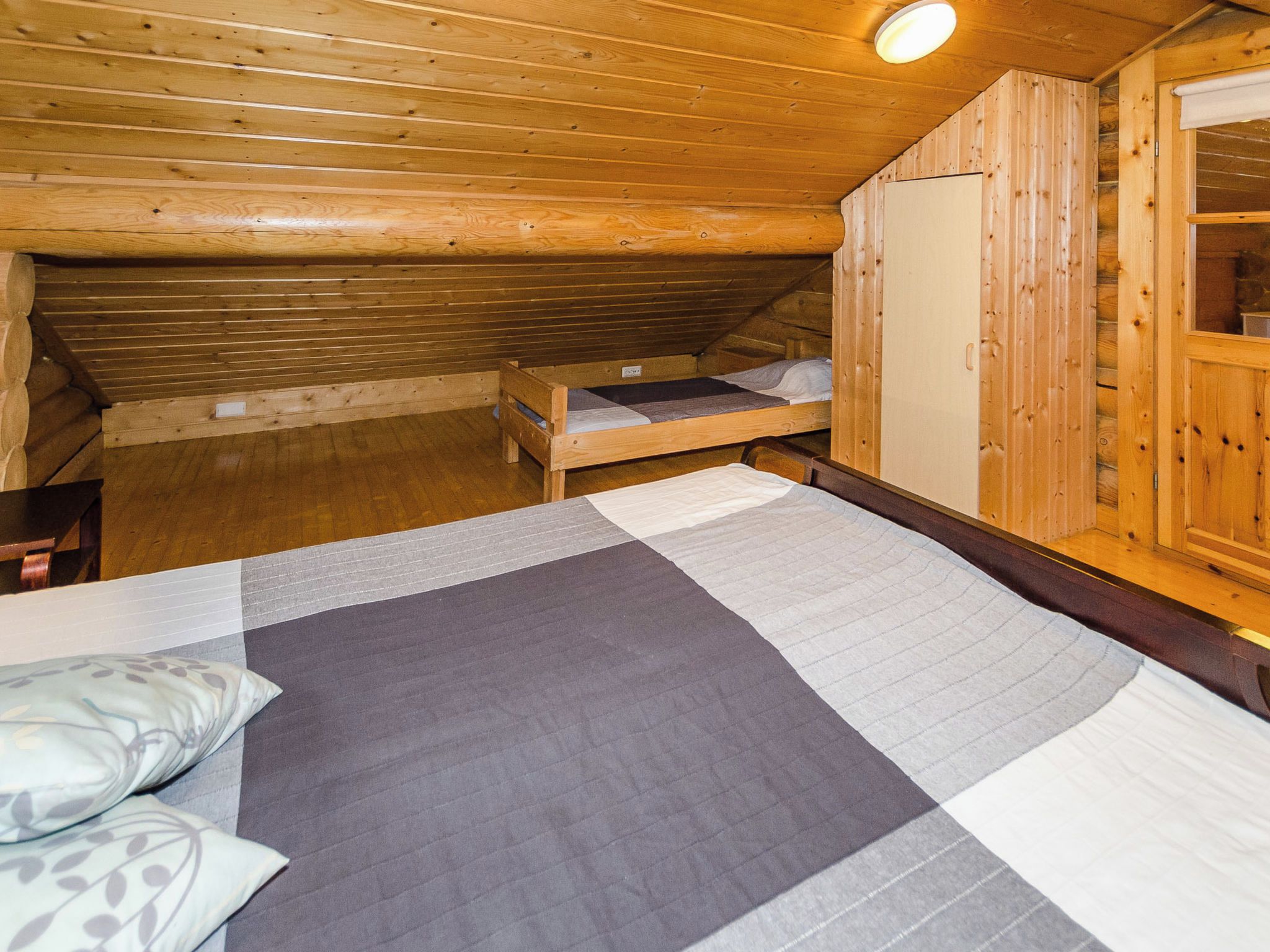 A small, cozy room with wooden walls and a sloped ceiling. There is a bunk bed with a mattress and pillows, a wooden wardrobe, and a small table with a chair.