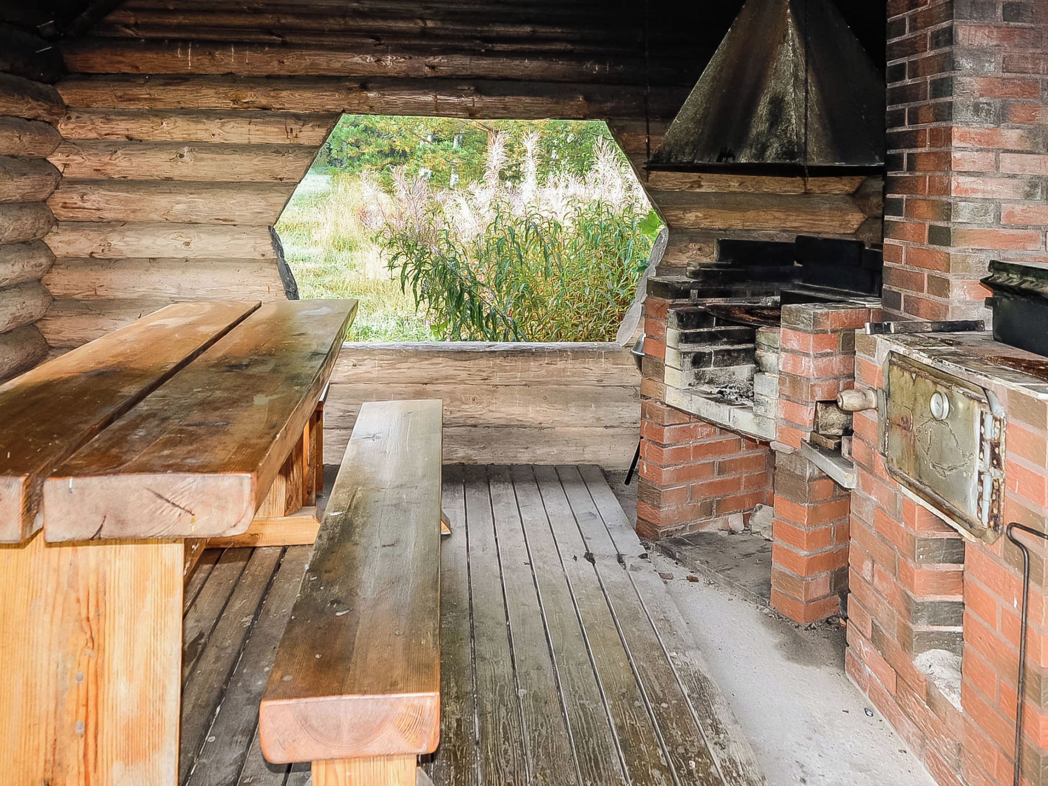 An outdoor wooden structure with a brick barbecue area, wooden benches, and a view of greenery.