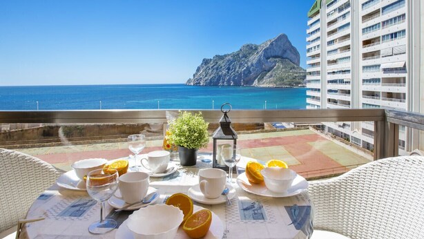 Une table de balcon agrémentée de tasses et d’assiettes blanches, ainsi que d’une plante en pot, d’où l’on peut admirer une vue sur la côte, avec une montagne et des bâtiments d’hôtel en arrière-plan.