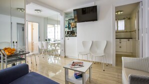 Un salon moderne avec un canapé, une table basse et un téléviseur fixé au mur. On y trouve également un coin repas avec des chaises et une table, ainsi qu'une cuisine visible à travers une porte ouverte.