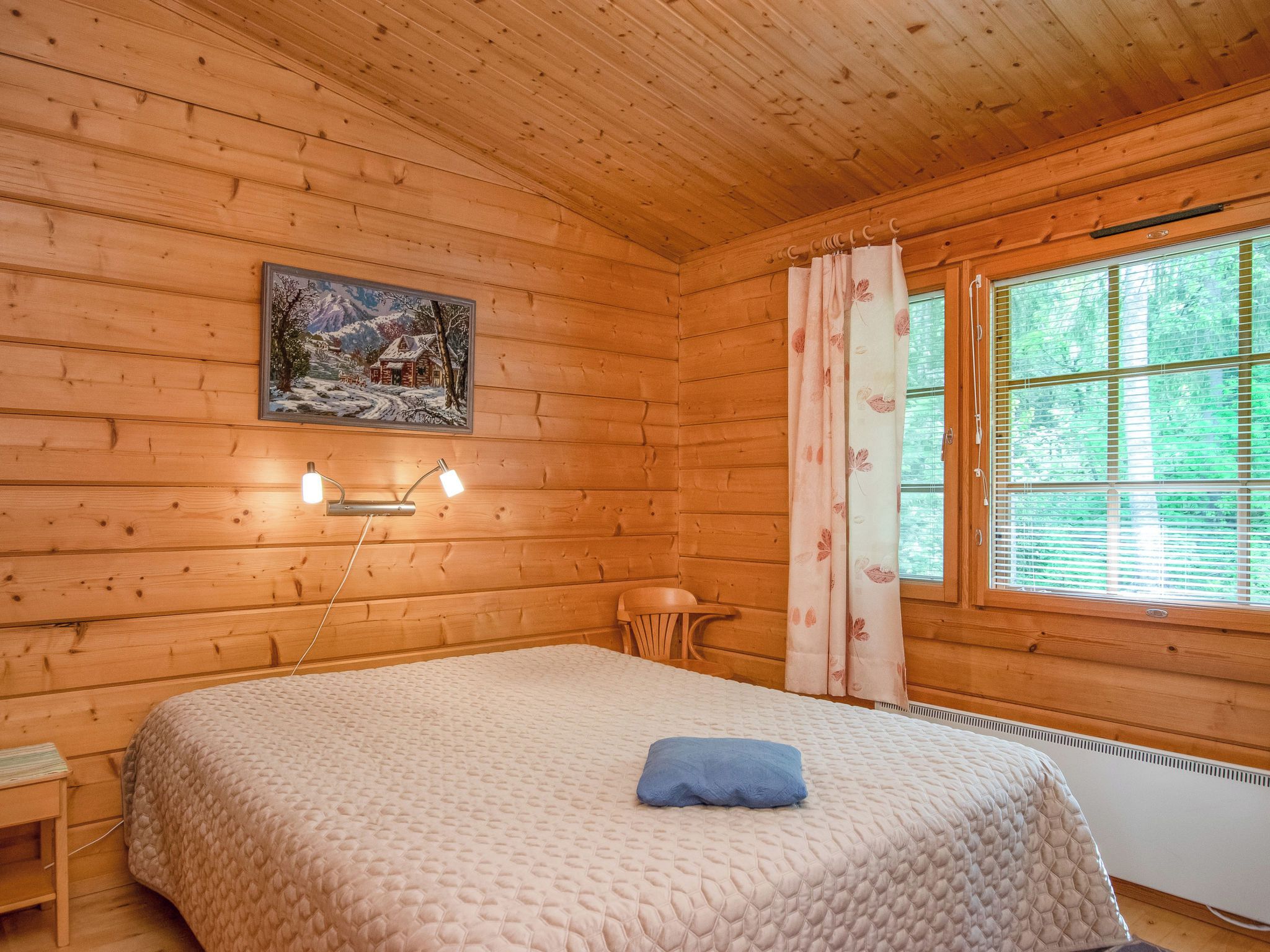 Ein gemütliches Blockhausszimmer mit einem Bett, einem kleinen Tisch, einem Sessel, einem Bild an der Wand und einem Fenster mit Vorhängen.