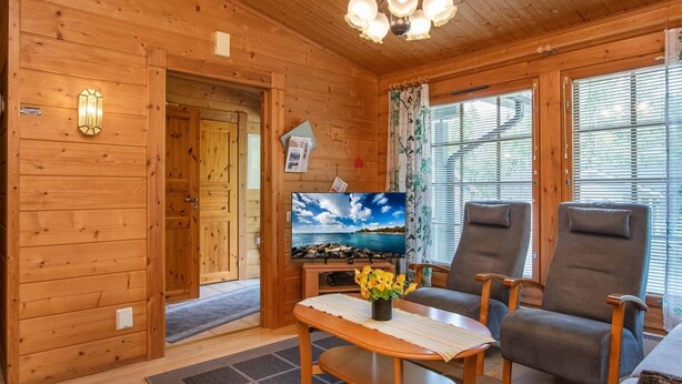 A wooden cabin interior with a television, two armchairs, a table with a vase of flowers, and a view of the sea through the window. - Rusakko by Interhome (Sotkamo)