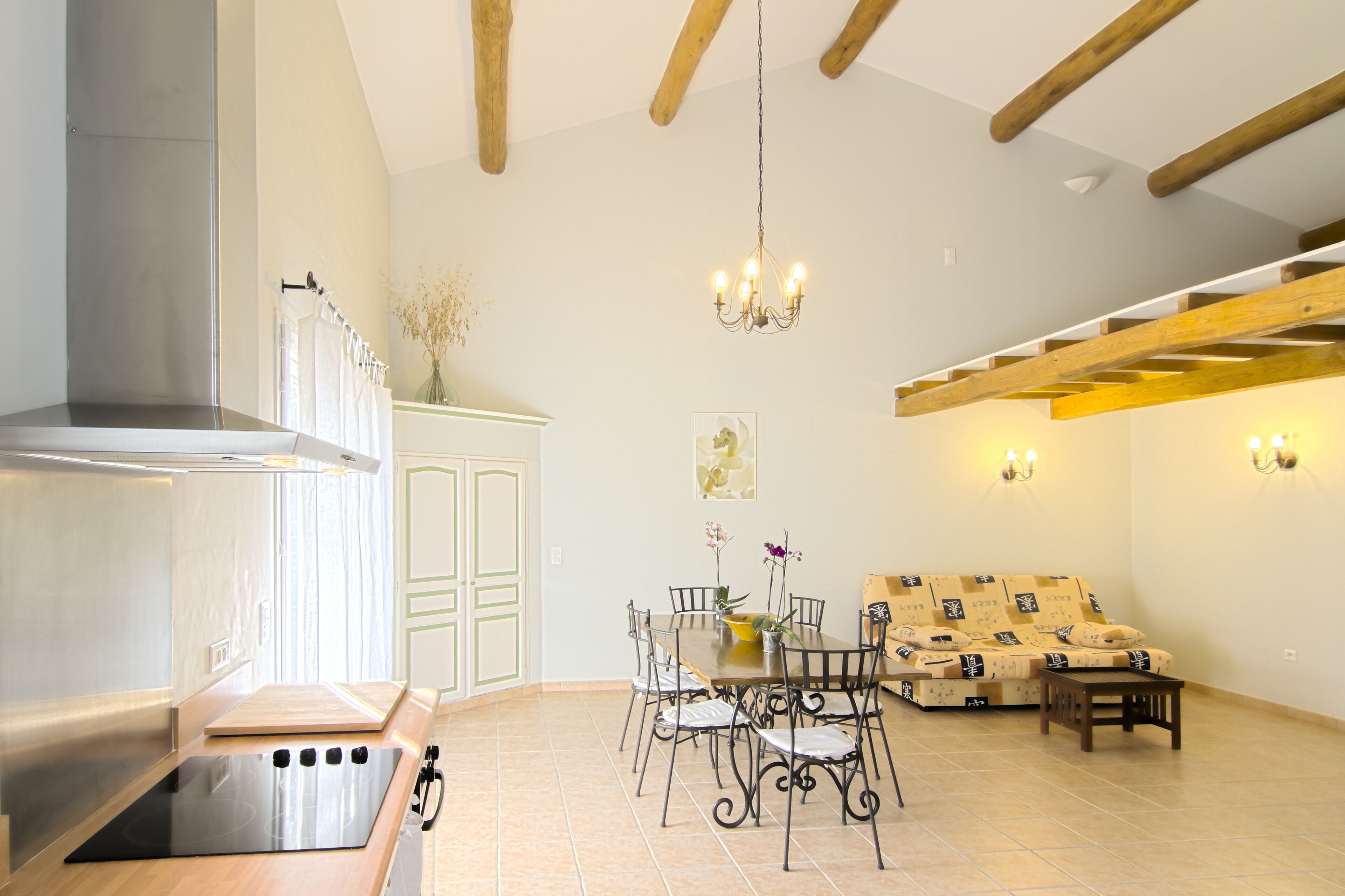 A modern kitchen with a built-in stove, dining table with chairs, and a sofa in a living area with exposed wooden beams and a chandelier.