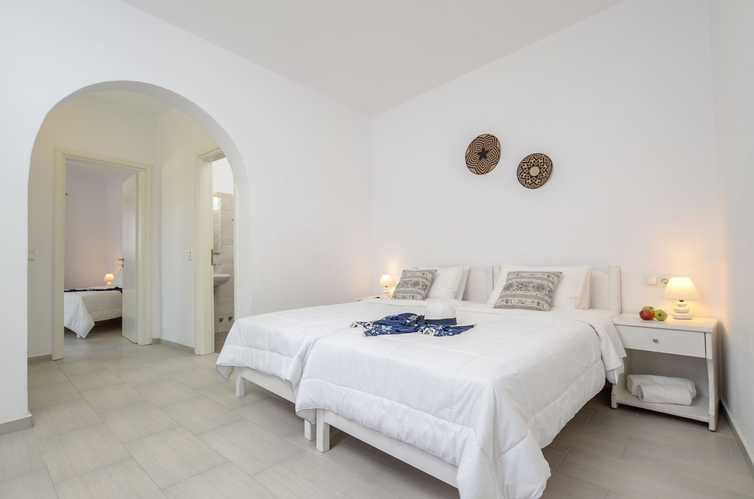 A bedroom with a large bed, two bedside lamps, a nightstand with fruit, and a mirror in the adjacent room.