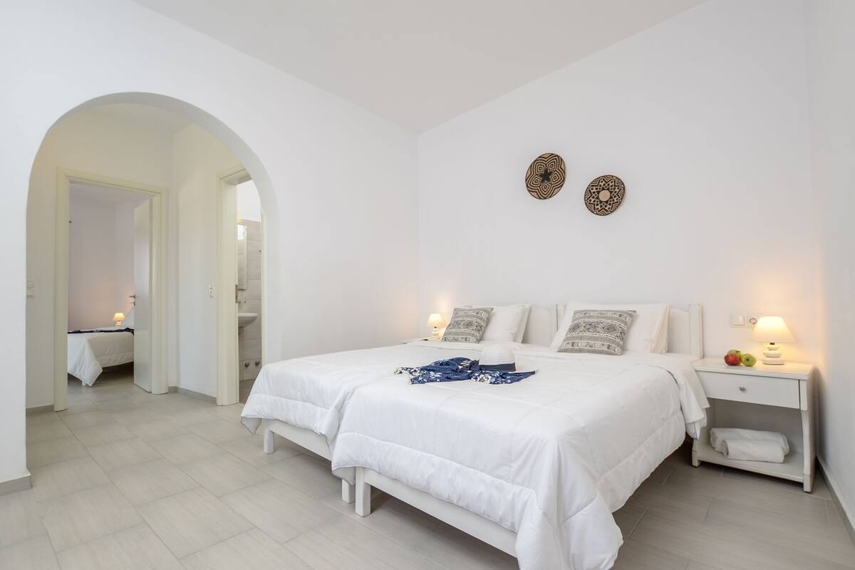 A bedroom with a large bed, two bedside lamps, a nightstand with fruit, and a mirror in the adjacent room.
