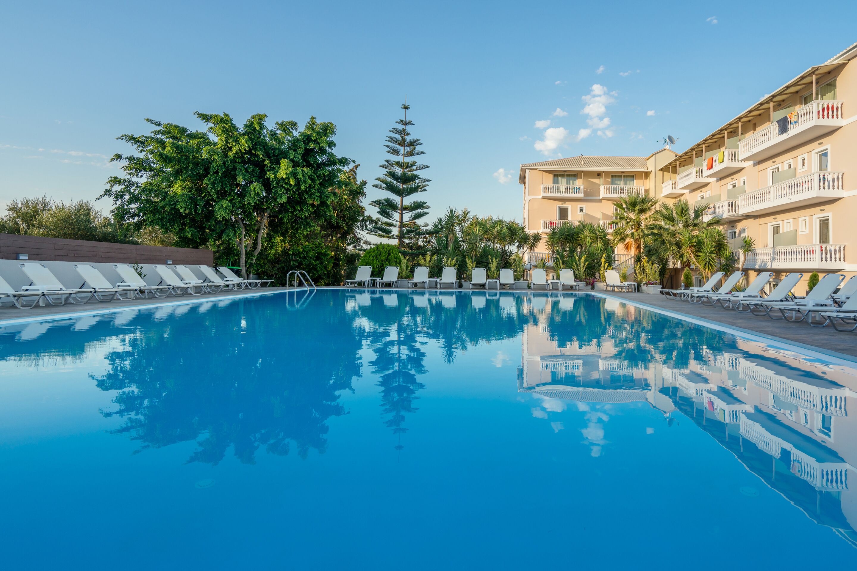 A swimming pool with lounge chairs, surrounded by trees and a multi-story building with balconies.