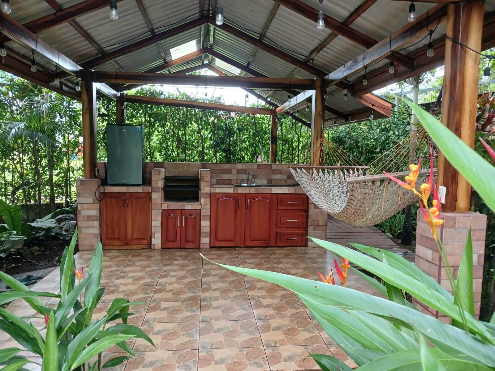 An outdoor kitchen with a grill, sink, and storage cabinets, surrounded by greenery and a hammock.