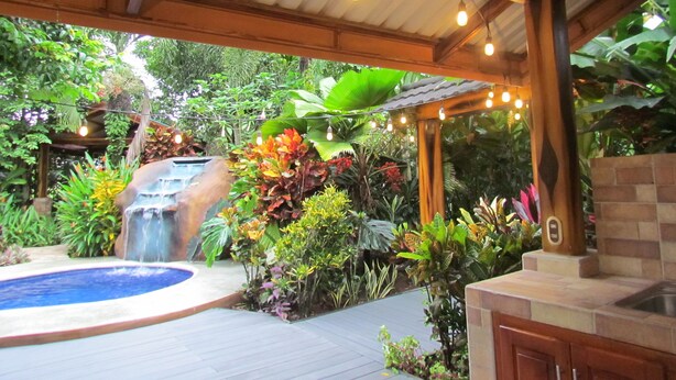 A covered outdoor area with a pool, waterfall, and various plants. - San Gabriel Apartments (La Fortuna)
