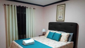 A bedroom with a bed, curtains, a nightstand, a lamp, and a framed picture on the wall. - San Gabriel Apartments (La Fortuna)