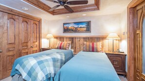 A bedroom with two beds, wooden wardrobe, ceiling fan, wall-mounted lamps, and a painting.