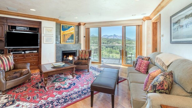 A living room with a fireplace, a television, and a large window offering a mountain view. - Penthouse At The Peaks 744 (Mountain Village)