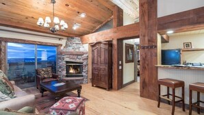 A spacious living area with a stone fireplace, wooden ceiling beams, and a large window overlooking a scenic view. - Beautiful 2 Bedroom Penthouse in Mountain Village, CO (Mountain Village)