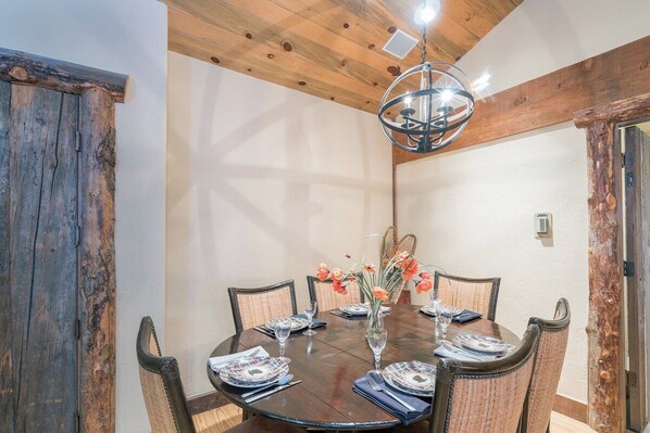 A dining area with a wooden table set for six, surrounded by wicker chairs, under a wooden ceiling with exposed beams.