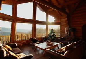A cozy log cabin interior with a large window, a Christmas tree, and a fireplace.