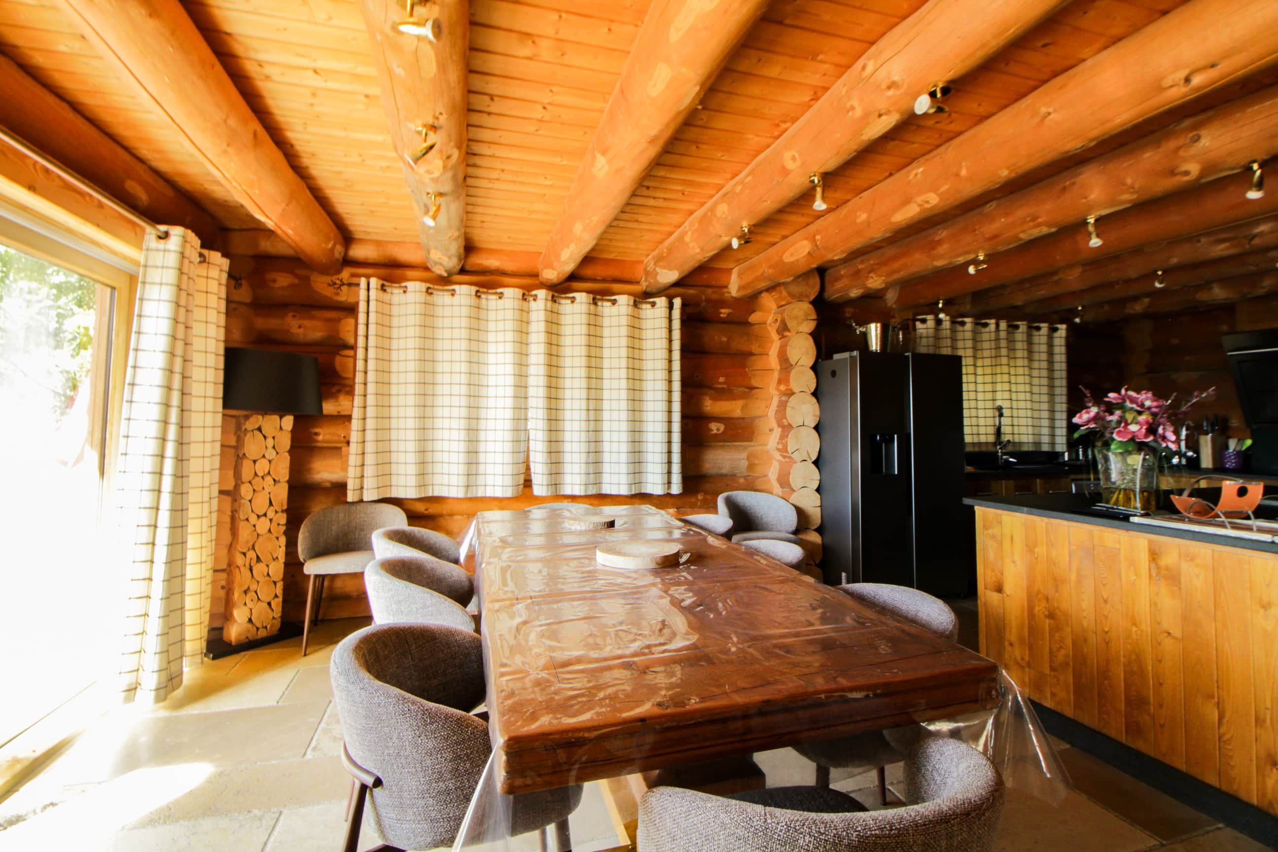 A wooden interior with a dining table, chairs, a kitchen area, and a window with curtains.