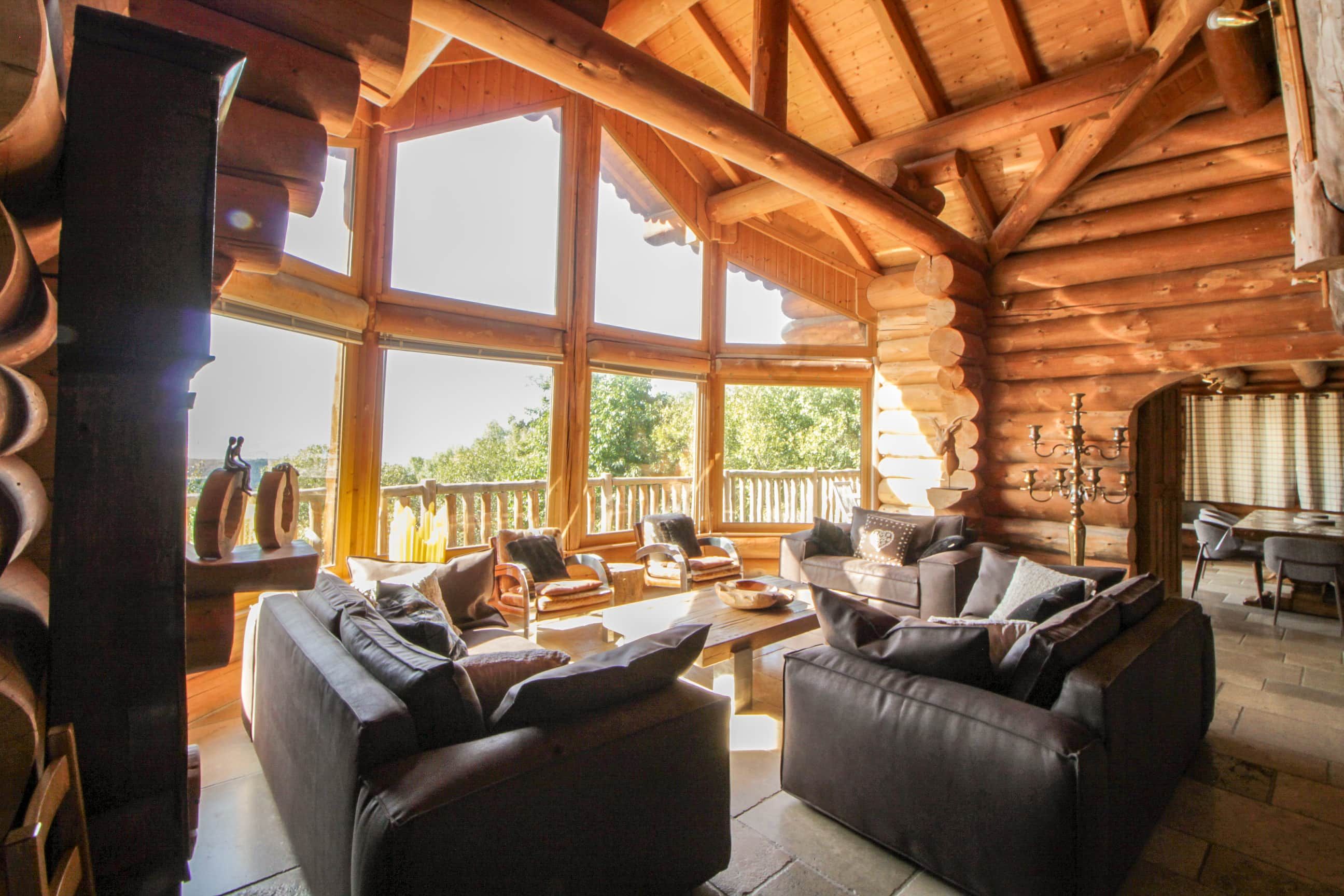 Interior d'una cabana de fusta amb una llar de foc, sofàs de pell i grans finestres que donen a l'exterior.