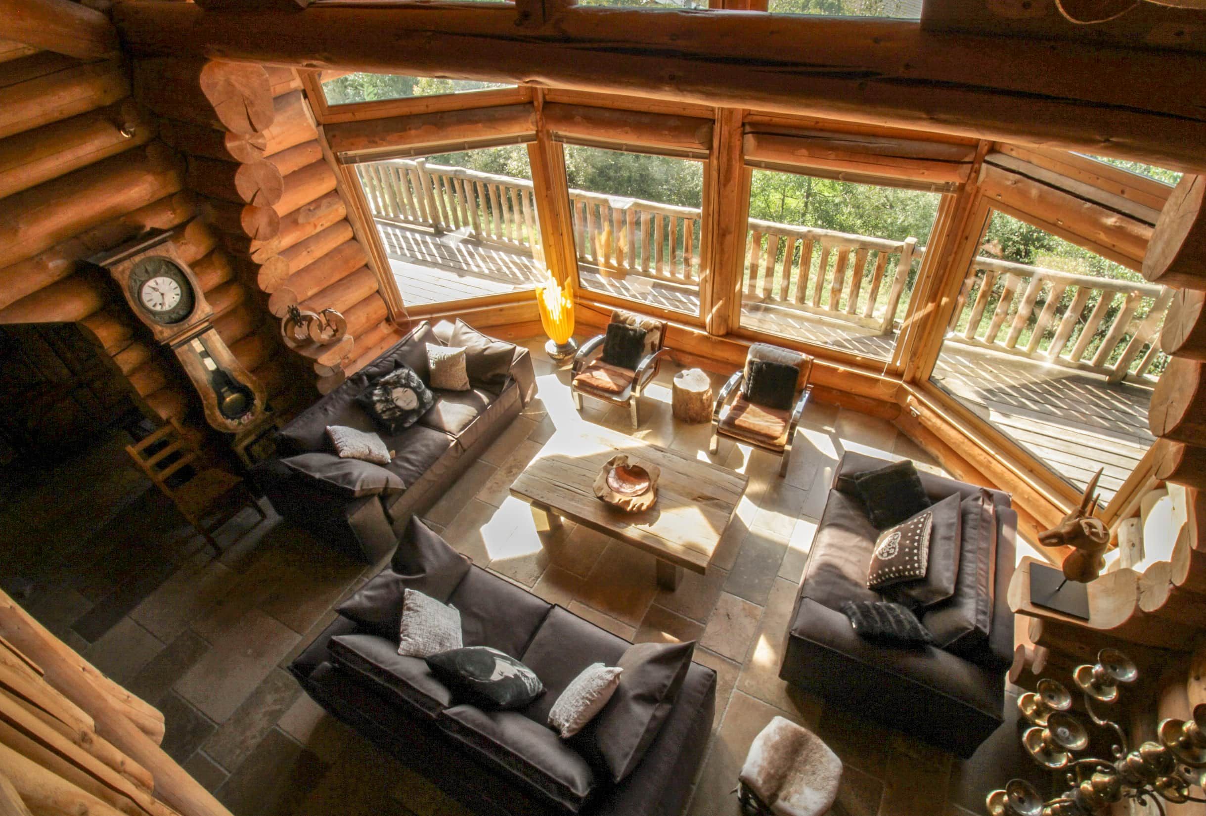 A log cabin interior with a fireplace, a sofa set, a coffee table, and a view of the outdoors through large windows.