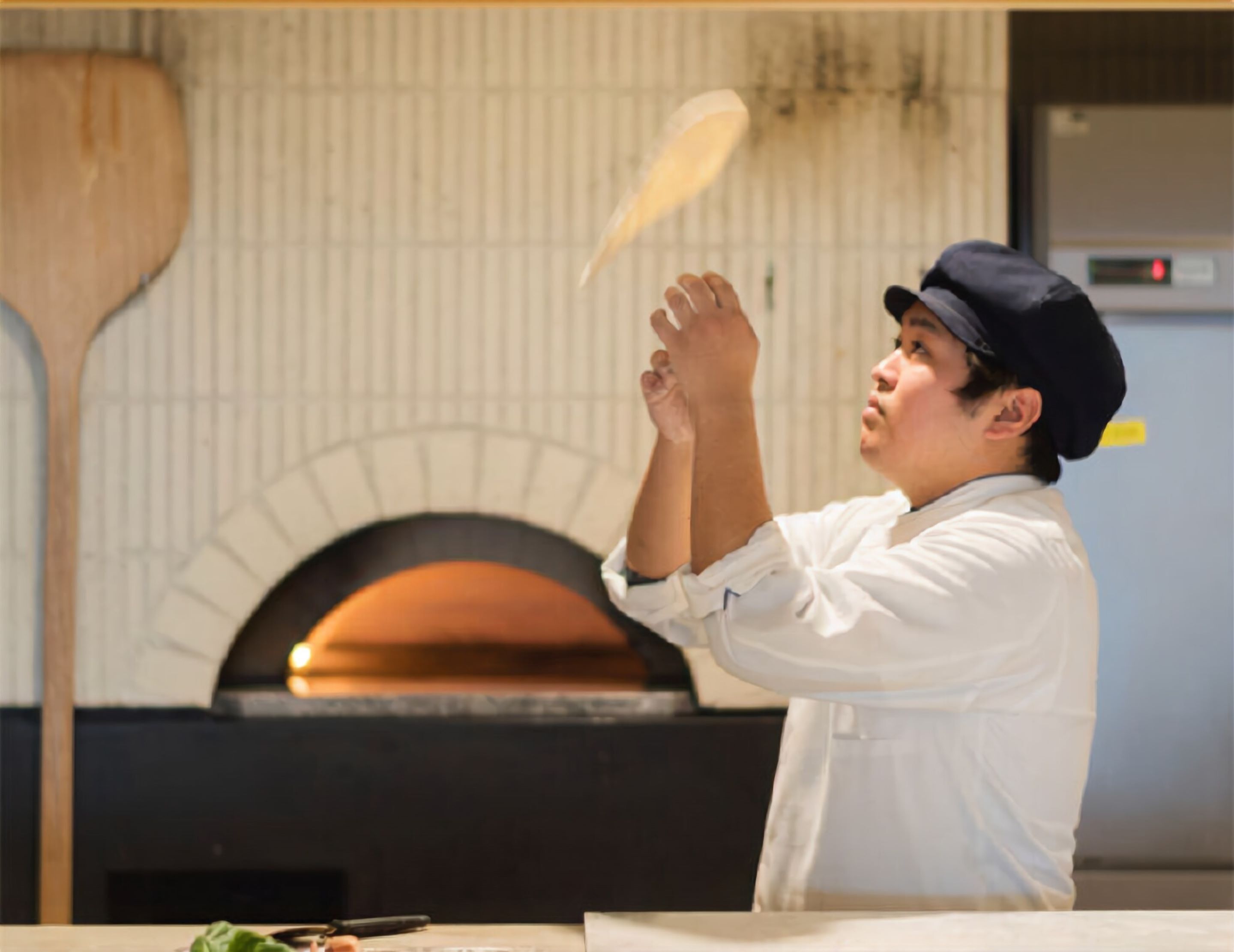 Une personne vêtue d’une chemise blanche et d’une casquette lance une pizza dans un four en briques.