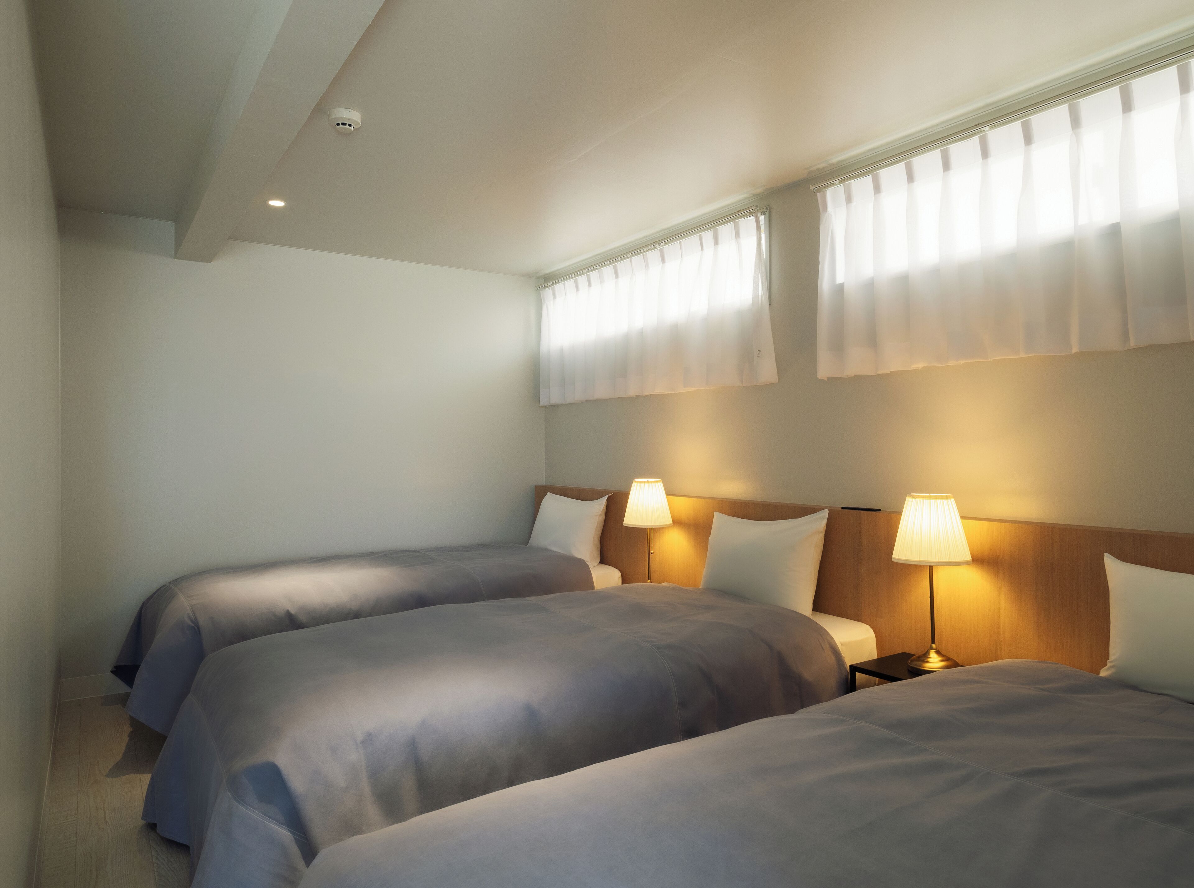 A hotel room with two beds, each with a bedside table and lamp. The room has a simple, clean design with white walls and a wooden headboard.