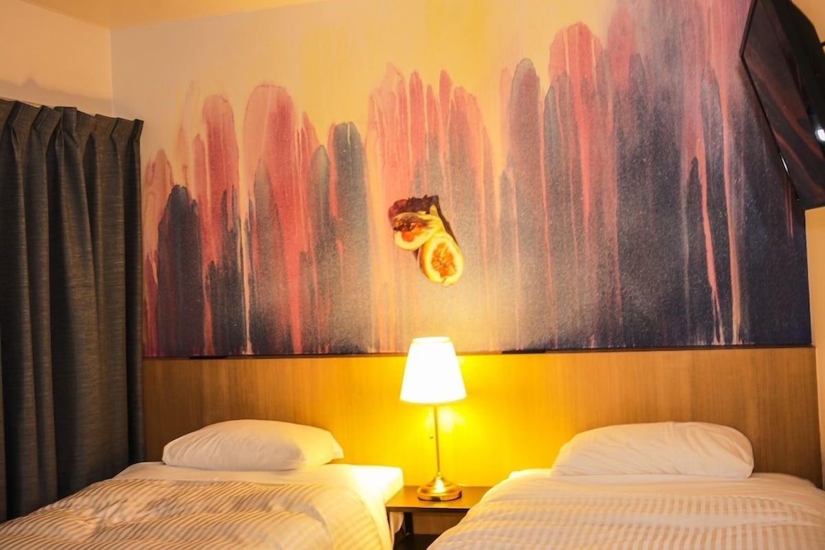 A hotel room with two beds, a nightstand with a lamp, and a large abstract painting on the wall.